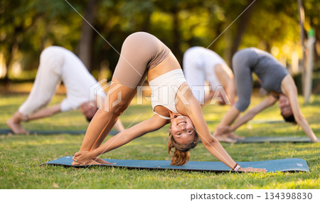 Yoga lesson visitors with coach perform Adho Mukha Svanasana in park 134398830