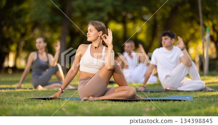 Girl performing Lord of Fishes Pose during group yoga practice outdoors Girl performing Lord of Fishes Pose during group yoga practice outdoors 134398845