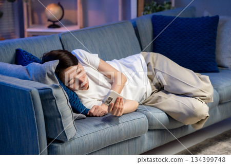 A woman lying on the sofa and looking at her smartphone A woman lying on the sofa and looking at her smartphone 134399748