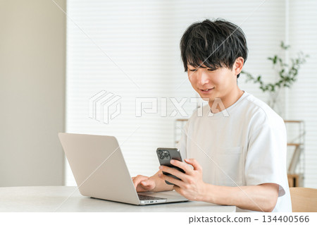 A young Asian man using a laptop and smartphone in the living room of his home (individual investor, investor, investment, asset management) 134400566