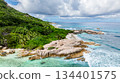 Waves break over granite rocks along a coastline fringed by lush greenery and clear waters. Seychelles, La Digue. 134401575