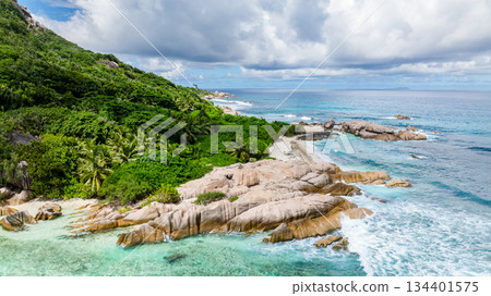Waves break over granite rocks along a coastline fringed by lush greenery and clear waters. Seychelles, La Digue. 134401575