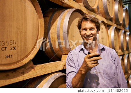 Portrait, smile and wine tasting with a man in the cellar of a distillery on a farm for the production of alcohol. Glass, industry and barrel with a happy male farmer drinking for quality control 134402623