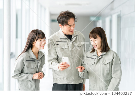 Male and female staff in the manufacturing, building, transportation and construction industries wearing work clothes talking while looking at their smartphones Male and female staff in the manufacturing, building, transportation and construction industries wearing work clothes talking while looking at their smartphones 134403257