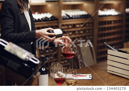 Woman in strict classical suite pours red wine from bottle to decanter, standing in cellar during sommelier exam certification. Woman in strict classical suite pours red wine from bottle to decanter, standing in cellar during sommelier exam certification. 134403278