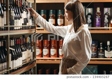 Indoor shot of experienced female sommelier taking wine bottle from shelf, choosing perfect variant for visitor based on his requests. Winemaking courses. Indoor shot of experienced female sommelier taking wine bottle from shelf, choosing perfect variant for visitor based on his requests. Winemaking courses. 134403287