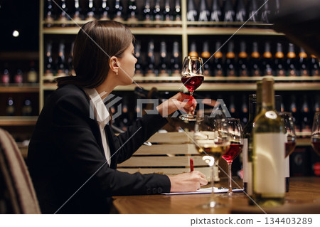 Experienced female sommelier tasting wine into glass sitting in cellar over plenty bottle shelf background. Experienced female sommelier tasting wine into glass sitting in cellar over plenty bottle shelf background. 134403289