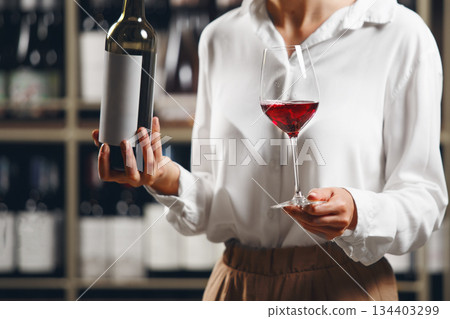 Close up woman hand hold bottle and glass with poured red wine on cellar background. 134403299