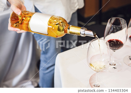 Pouring White Wine From Bottle to the Glass Near Red Wine Standing on Table Pouring White Wine From Bottle to the Glass Near Red Wine Standing on Table 134403304
