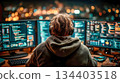 A man in a hoodie and headphones sits in front of several glowing monitors. A programmer at work. City lights in the background 134403518
