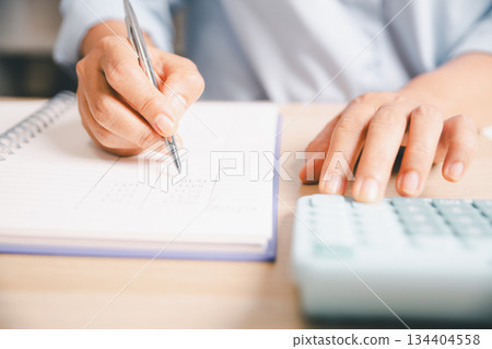 Close-up of office worker writing financial figures in notebook while using a calculator, illustrating budgeting, accounting tasks, expense tracking, professional financial management in workplace. 134404558