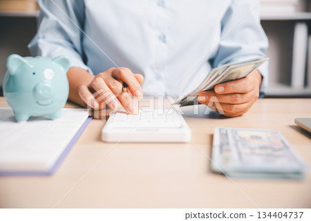 Office worker counting cash while using a calculator beside a piggy bank, illustrating personal finance, saving money, budgeting, expense tracking and financial management in modern workspace. 134404737
