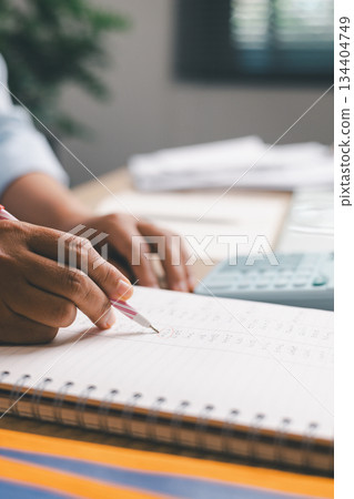 Close-up of office worker marking, writing financial figures in notebook, illustrating accounting tasks, record keeping, expense tracking, budgeting, professional financial management in workplace. 134404749