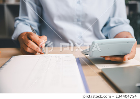 Close-up of office worker writing financial records in notebook while holding calculator, illustrating accounting tasks, expense tracking, budgeting and professional financial management in workplace Close-up of office worker writing financial records in notebook while holding calculator, illustrating accounting tasks, expense tracking, budgeting and professional financial management in workplace 134404754