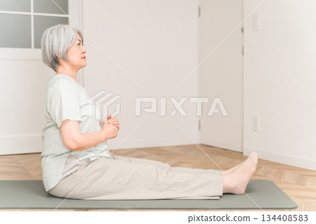 A senior woman with gray hair stretches and does flexibility exercises on a yoga mat in her room. A senior woman with gray hair stretches and does flexibility exercises on a yoga mat in her room. 134408583