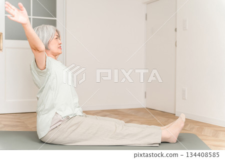 A senior woman with gray hair stretches and does flexibility exercises on a yoga mat in her room. 134408585