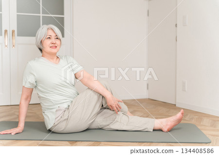 A gray-haired senior woman stretching and doing flexibility exercises on a yoga mat in her room 134408586