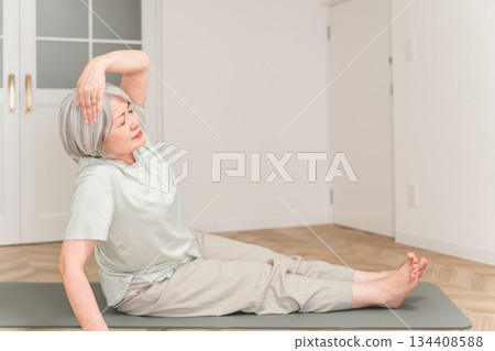 A gray-haired senior woman stretching and doing flexibility exercises on a yoga mat in her room 134408588