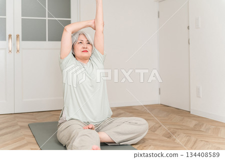 一位頭髮花白的老婦人在房間裡的瑜珈墊上做伸展運動和柔軟度練習。 134408589