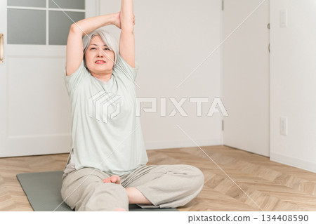 A gray-haired senior woman stretching and doing flexibility exercises on a yoga mat in her room 134408590