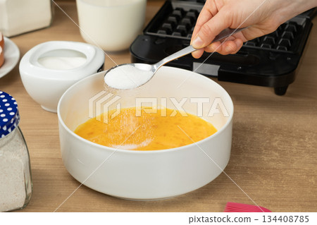 Close-up of a Hand Adding Sugar with a Spoon into a Bowl of Whisked Eggs for Baking Waffles Close-up of a Hand Adding Sugar with a Spoon into a Bowl of Whisked Eggs for Baking Waffles 134408785