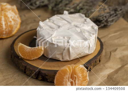 fresh white soft cheese with white mold lies on a wooden board on crumpled yellow paper, soft round cheese and juicy sweet orange tangerines 134409508