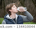 Athlete and sporty caucasian man drinking water from bottle at public park after workout. Outdoors. Water balance. Young Man Training Outdoors 134410044