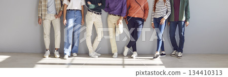 Diverse group's lower half of seven people dressed in casual clothes standing against white wall. 134410313