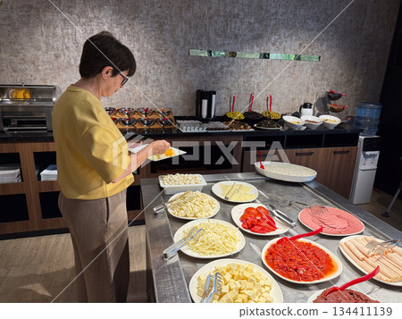 Woman selecting food from hotel breakfast buffet with cheese and cold cuts. Morning routine, self service dining, travel lifestyle, and everyday meal choice in restaurant interior. Woman selecting food from hotel breakfast buffet with cheese and cold cuts. Morning routine, self service dining, travel lifestyle, and everyday meal choice in restaurant interior. 134411139