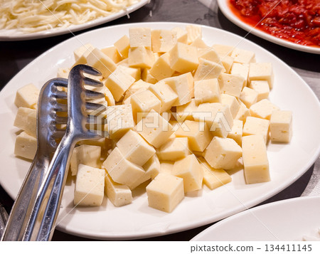 Cubed yellow cheese served on plate with metal tongs at breakfast buffet. Dairy product selection, protein nutrition, self service catering, and hotel dining experience. 134411145