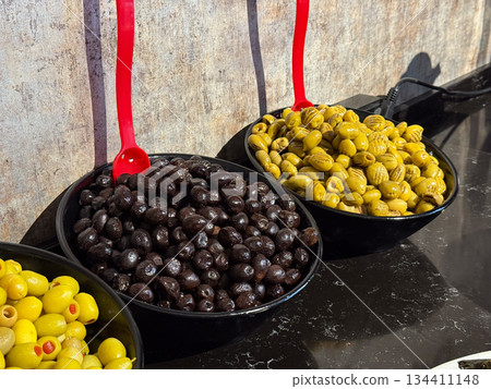 Black and green olives served in bowls with red ladles on buffet. Savory food assortment, Mediterranean flavors, catering service, and self service dining experience. Black and green olives served in bowls with red ladles on buffet. Savory food assortment, Mediterranean flavors, catering service, and self service dining experience. 134411148