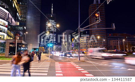 Warsaw, Poland. 4 November 2025. Blurred people cross a street at a crosswalk in center city in the night. Traffic lights glowing, and buildings towering around them. Nearby with cars passing. 134411587