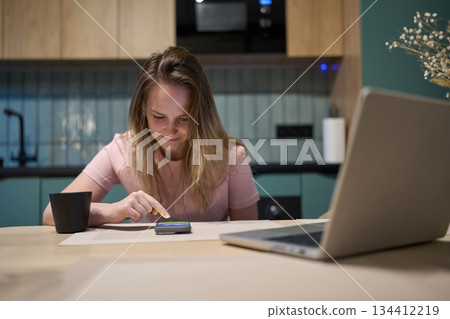 Young woman using smartphone at table with laptop in modern kitchen 134412219