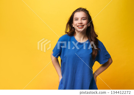 Positive teen girl with dental braces in blue t-shirt. Hands behind head, wide smile, looks at camera on yellow backdrop. Dental care services, orthodontic clinics, teen health campaigns, youth 134412272