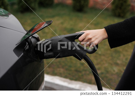 Unrecognisable woman inserting EV charger plug from public or home charging station for modern electric or hybrid car at special place for e-car. Concept eco-friendly sustainability energy car 134412291