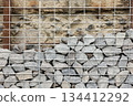 Stone fence in steel mesh and green lawn. Close up of gabion with tightly packed stones of varying sizes and textures. Combination of natural materials and industrial design. 134412292