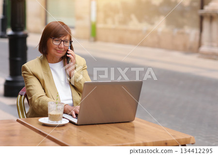 Mature office employee woman talks on smartphone with smile about upcoming business trip while sitting on terrace in cafe with cup of coffee and laptop, happy businesswoman working remotely online. 134412295