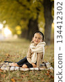 Adorable little child girl 6-7 years old smiling looks at camera, sits on plaid at outdoors at autumn park. Children's day. Stylish kid wears beige sweater and black headband. Happy childhood. 134412302