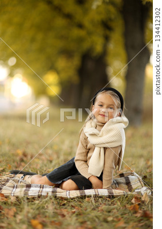 Adorable little child girl 6-7 years old smiling looks at camera, sits on plaid at outdoors at autumn park. Children's day. Stylish kid wears beige sweater and black headband. Happy childhood. 134412302