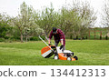 Man in casual clothes takes out bag with cut grass of lawn mower after mowing lawn with at own backyard. Husband takes care of garden. Modern gasoline garden equipment. Landscaping work at home 134412313
