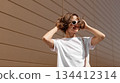 Attractive smiling young woman in fashionable sunglasses enjoys sunlights outdoors on warm summer day. Happy stylish girl with short curly hair in good mood. Concept of youth urban lifestyle 134412314