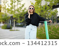 Cheerful woman in sunglasses near electric scooter or e-scooter talks on smartphone, at modern residential district on summer day. Concept of urban eco-friendly lifestyle and sustainability living 134412315