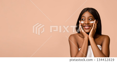 Beautiful african american young lady with face cream or serum on her cheeks. Smiling topless black woman with eye cream on her face, beige studio background, panorama with copy space Beautiful african american young lady with face cream or serum on her cheeks. Smiling topless black woman with eye cream on her face, beige studio background, panorama with copy space 134412819