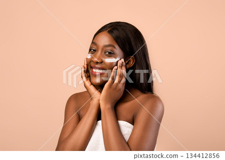 Nude black woman applying white eye cream after shower, beige studio background, closeup. Smiling beautiful african american lady wrapped in towel rubbing under-eyes zone with serum or cream 134412856