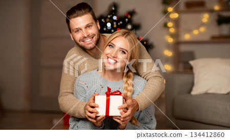 Cheerful young couple hugging and holding gift box, exchanging with Christmas presents at home, empty space. Smiling man and woman presenting xmas gifts to each other, celebrating winter holidays 134412860