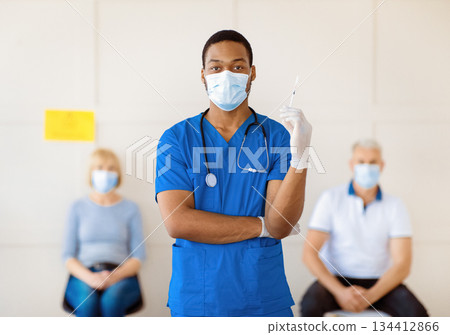 Portrait of young African American doctor in mask and blue uniform holding syringe with covid-19 vaccine at health centre, patients waiting for coronavirus immunization. Epidemic prevention measures Portrait of young African American doctor in mask and blue uniform holding syringe with covid-19 vaccine at health centre, patients waiting for coronavirus immunization. Epidemic prevention measures 134412866
