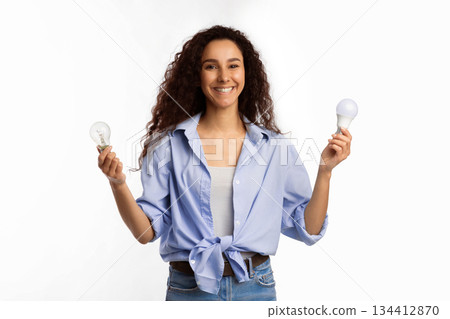 Save Energy. Cheerful Lady Holding Two Lightbulbs Choosing Energy Saving Light Bulb Standing Over White Studio Background, Looking At Camera. Safe Light-Bulbs Disposal Service Concept Save Energy. Cheerful Lady Holding Two Lightbulbs Choosing Energy Saving Light Bulb Standing Over White Studio Background, Looking At Camera. Safe Light-Bulbs Disposal Service Concept 134412870