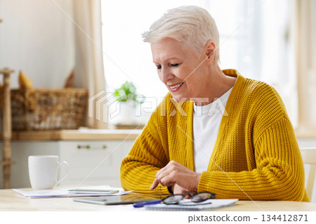 Positive grey-haired senior woman using digital tablet in kitchen, taking notes, counting month spending, copy space. Smiling old lady grandmother enjoying nice mobile application Positive grey-haired senior woman using digital tablet in kitchen, taking notes, counting month spending, copy space. Smiling old lady grandmother enjoying nice mobile application 134412871