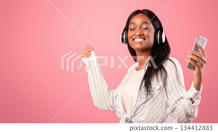 Lovely black lady with headphones and mobile device listening to music and dancing on pink studio background. Beautiful African American woman moving to her favorite song with closed eyes Lovely black lady with headphones and mobile device listening to music and dancing on pink studio background. Beautiful African American woman moving to her favorite song with closed eyes 134412883
