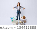 Happy young lady sorting waste, pointing at containers with different kind of garbage, millennial eco-volunteer woman enjoying trash recycling, standing over light studio background, full-length shot 134412888
